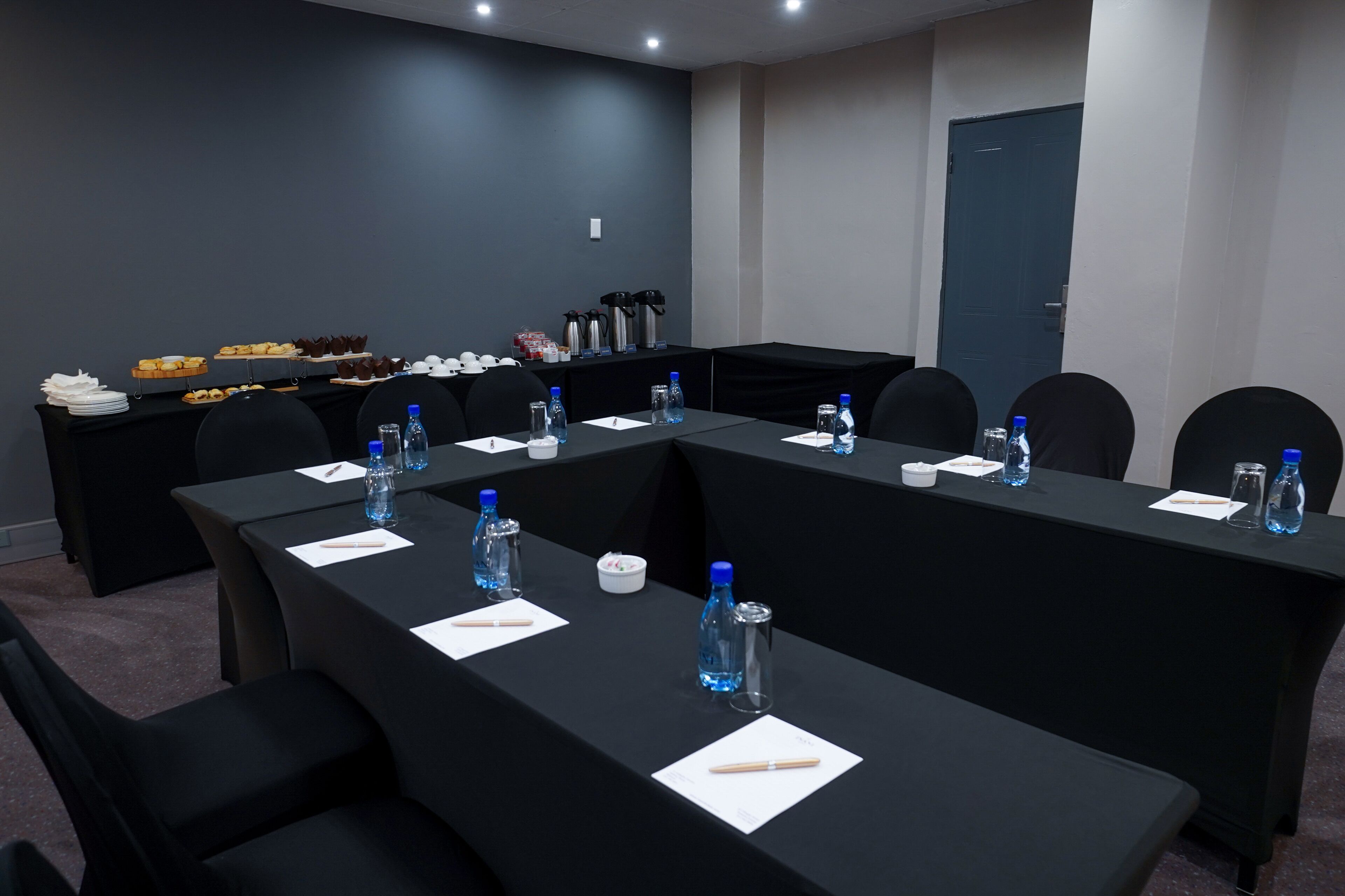 A conference room with a long table, chairs, water bottles, and a buffet table with various food items.