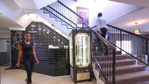 A hotel lobby with a staircase, a reception area with a glass display case, and a black brick wall with a coffee cup design. - The Gallagher Hotel (Midrand)
