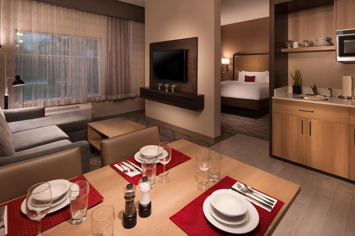 Une chambre d’hôtel équipée d’une table à manger pour quatre personnes, d’un téléviseur fixé au mur, d’un lit avec une tête de lit et d’une kitchenette.
