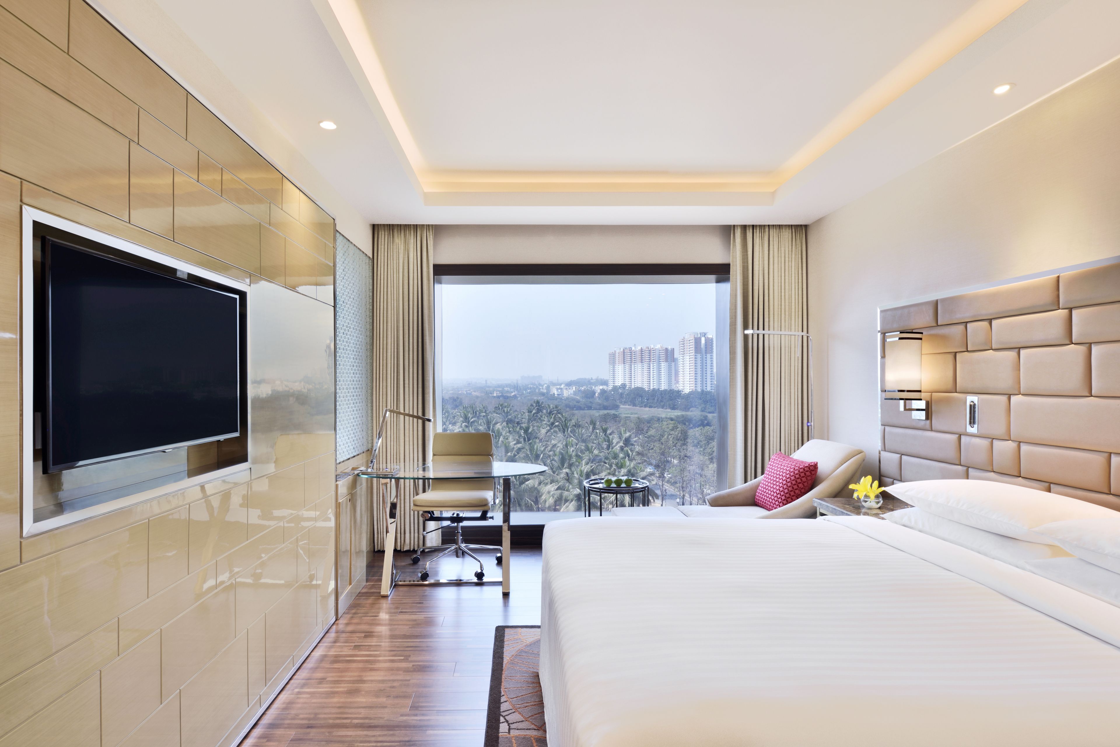 Une chambre d’hôtel moderne dotée d’un grand lit, d’un téléviseur à écran plat fixé au mur, d’une petite table avec une chaise et offrant une vue sur le paysage urbain par le biais d’une grande fenêtre.
