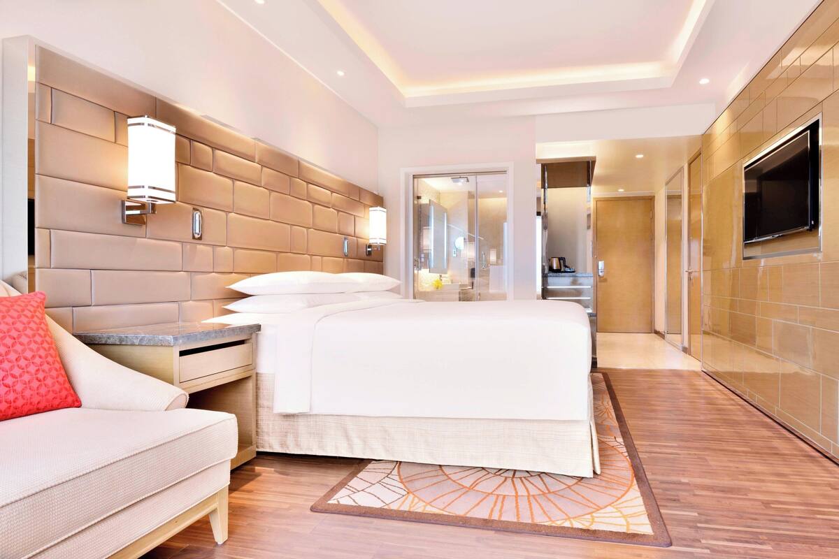 Une chambre d’hôtel moderne dotée d’un grand lit, d’un canapé, d’une télévision et d’une salle de bain visible à travers une porte en verre.