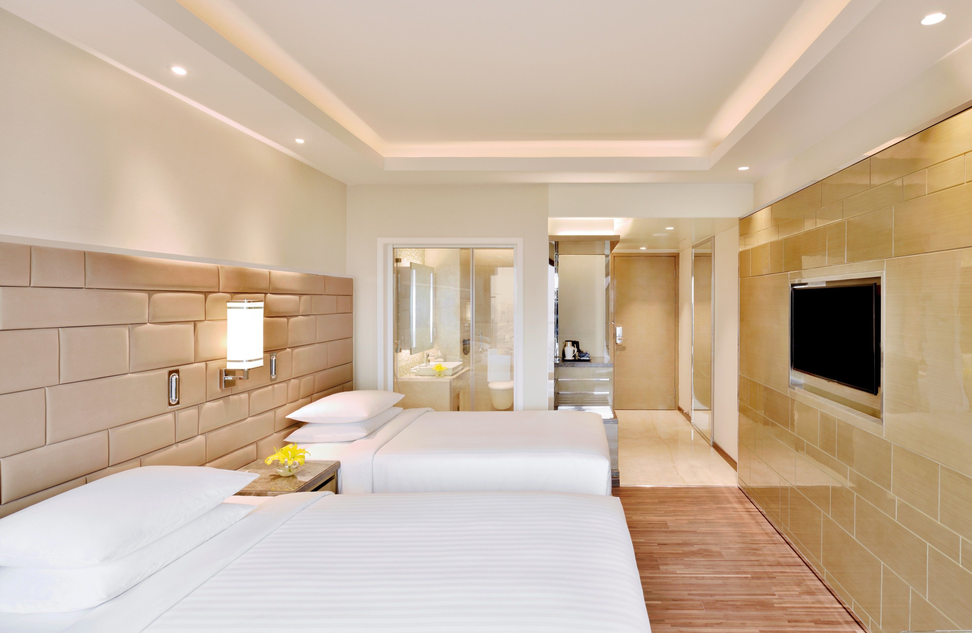 A hotel room with two beds, a TV, a bathroom, and a hallway leading to a kitchen.