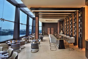 Un restaurante moderno con una barra larga, mesas redondas y asientos de bar. Amplias ventanas ofrecen vistas de la ciudad.