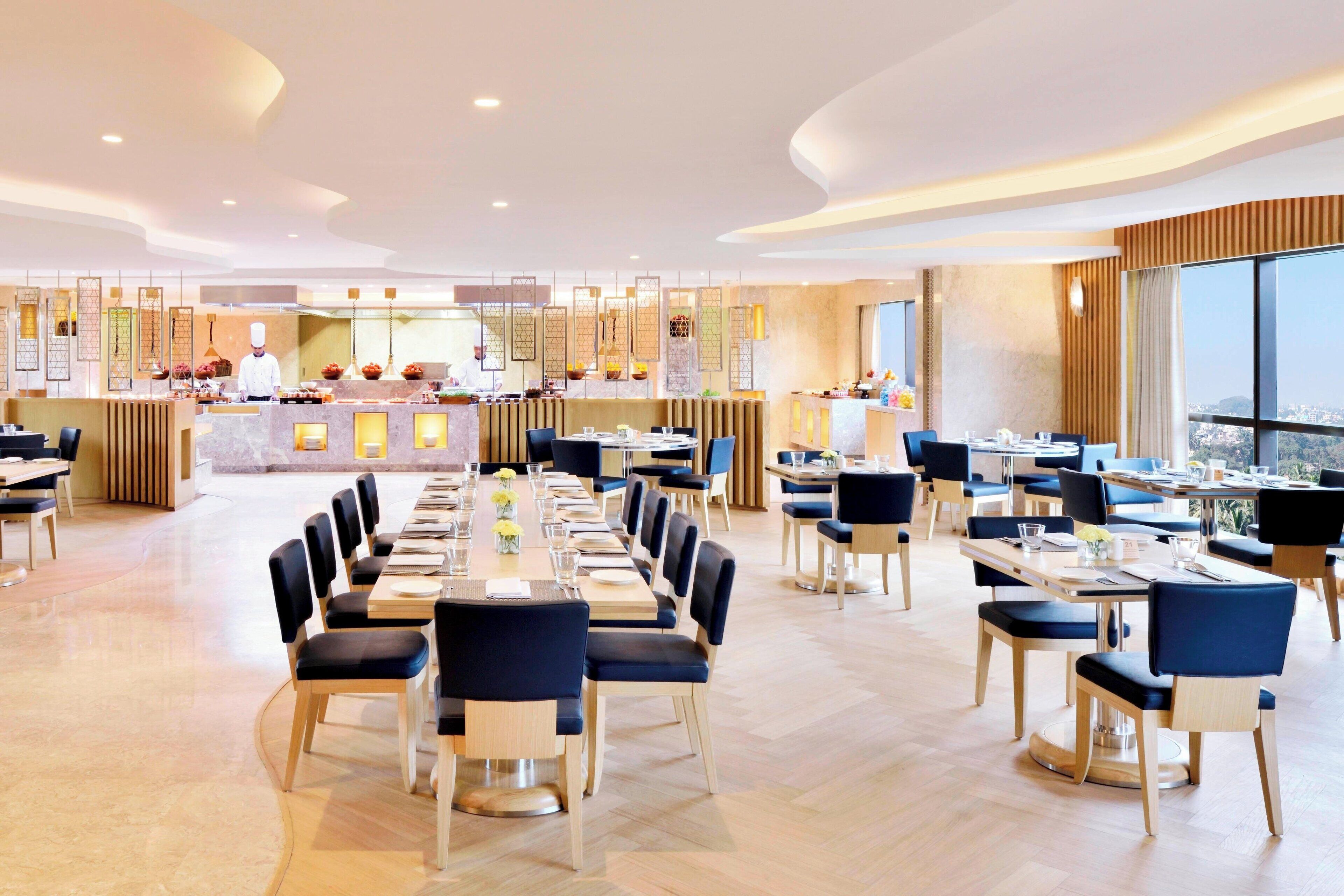 Un grand espace dédié à la salle à manger, avec plusieurs tables et chaises, une cuisine centrale et de grandes fenêtres offrant une vue dégagée.