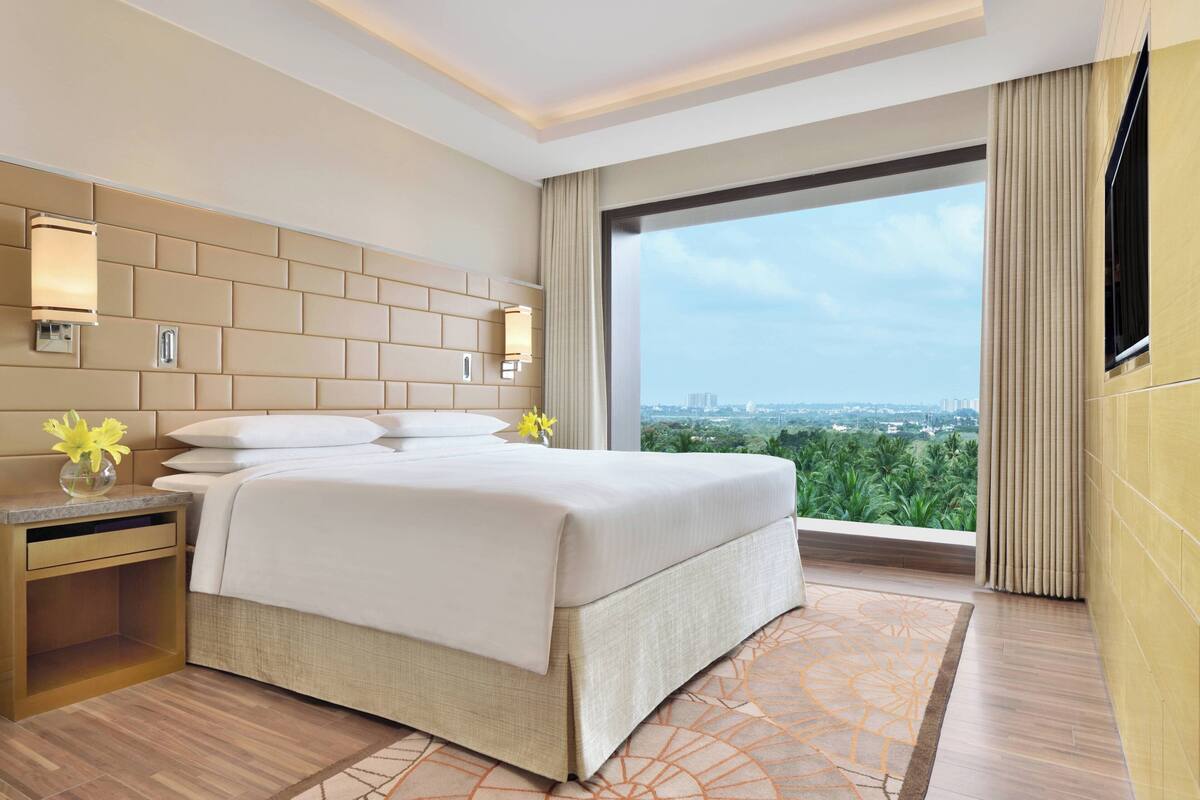 Une chambre d’hôtel moderne avec un grand lit, une table de chevet, un vase de fleurs et une vue sur la ville par une grande fenêtre.
