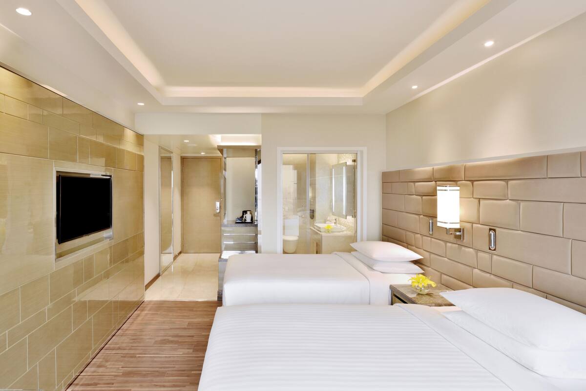 Une chambre d’hôtel avec deux lits, une télévision, un miroir et une salle de bain visible à travers une porte en verre.