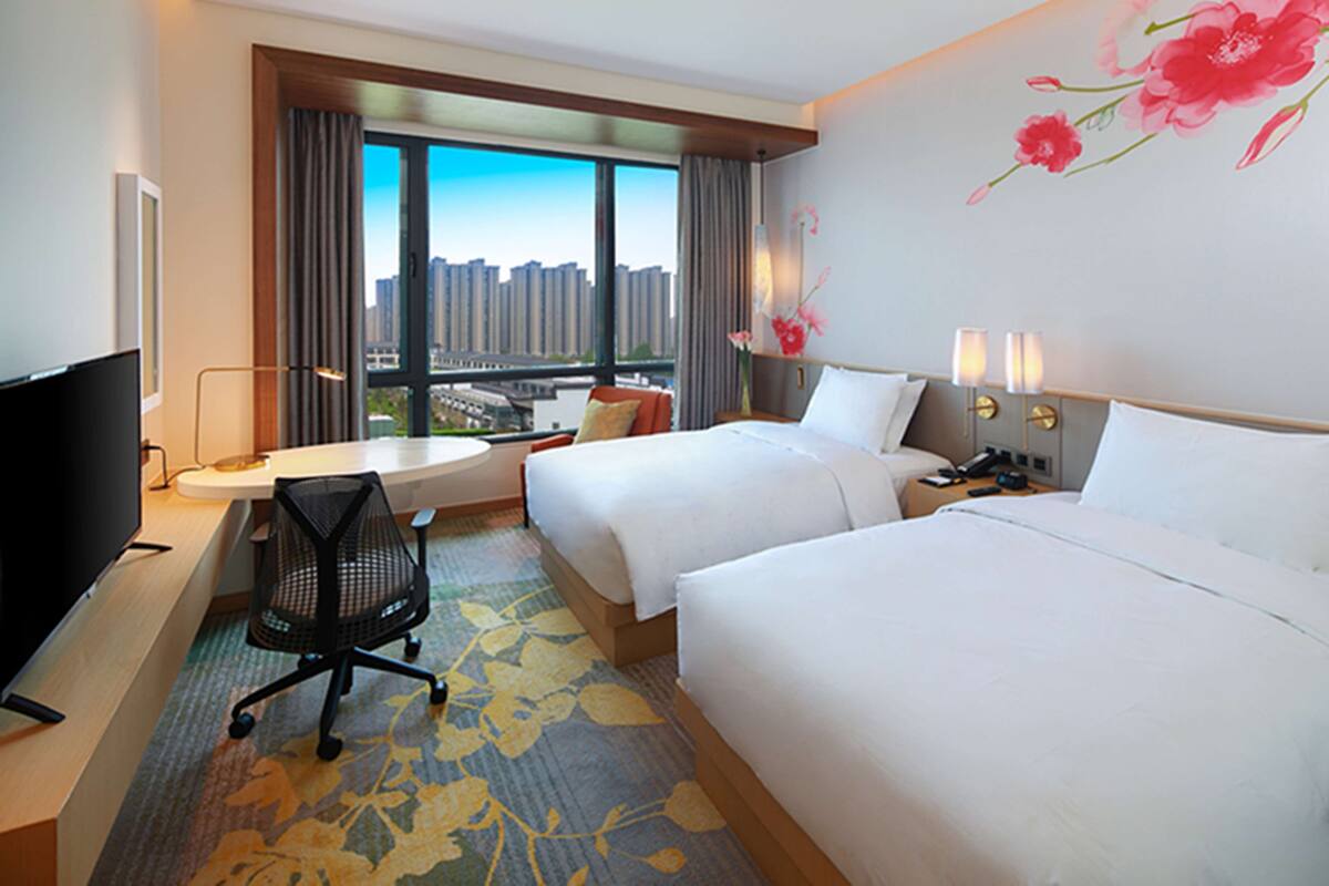 A hotel room with two beds, a desk, a TV, and a large window with a city view.