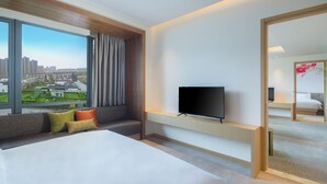 Une chambre d’hôtel avec un grand lit, un téléviseur posé sur un meuble, une fenêtre donnant sur un paysage urbain et une porte coulissante menant à une autre pièce.