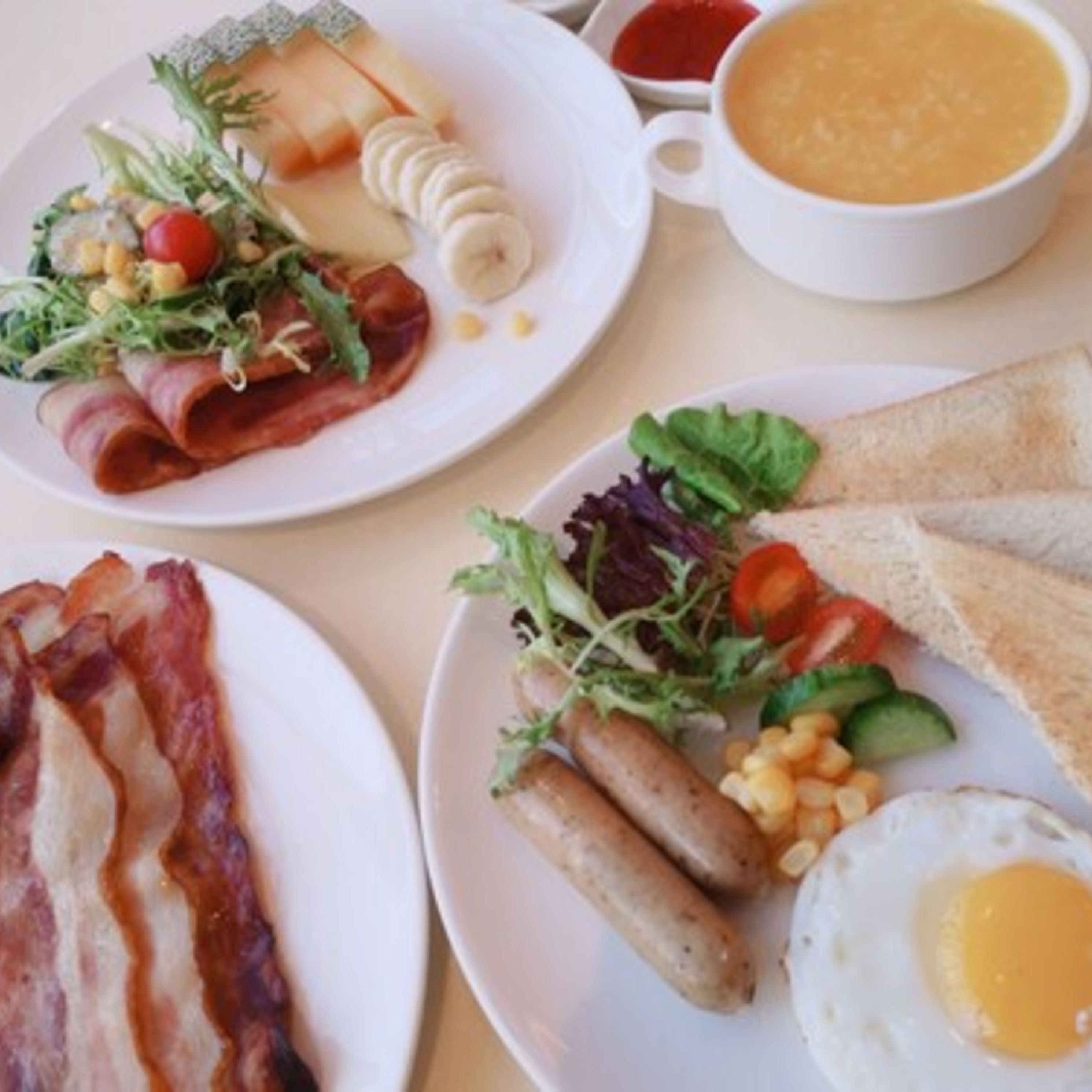 A breakfast setting with plates of bacon, eggs, sausages, and toast, along with a cup of soup and a salad.