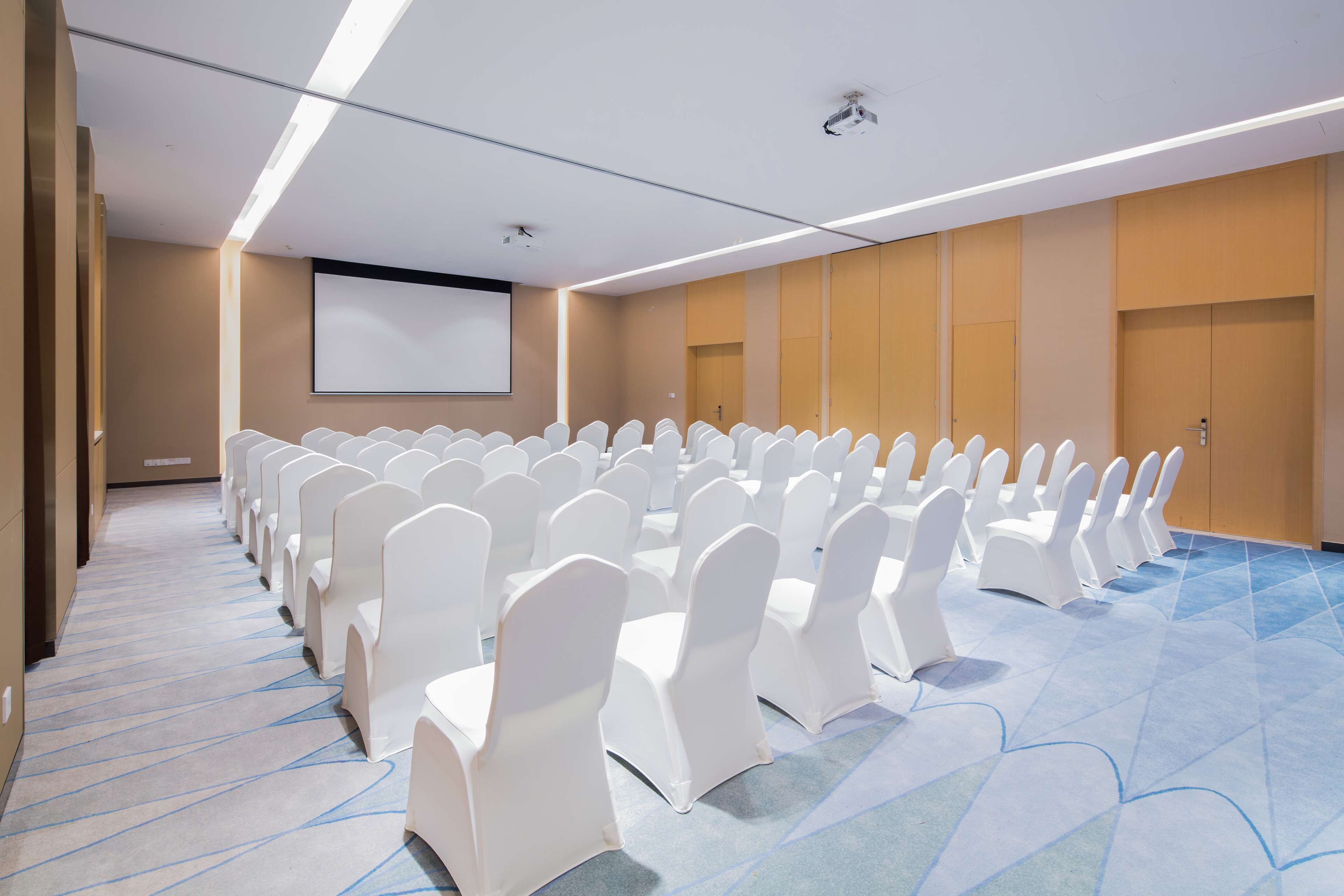 A conference room with rows of white chairs and a large screen on the wall.