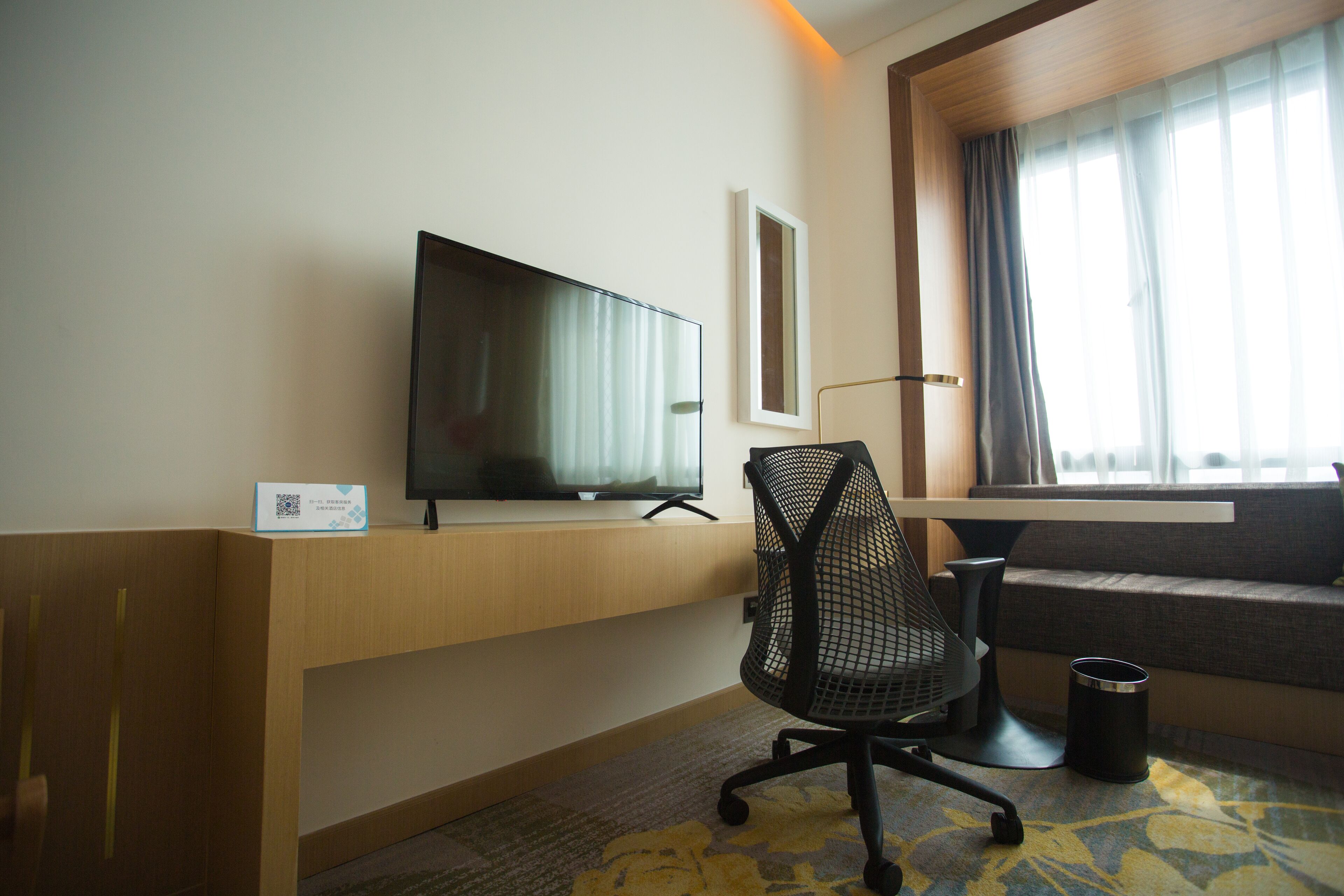 Hotellihuone, jossa on litteänäyttöinen televisio puisen jalustan päällä, musta verkkomainen toimistotuoli, pieni roskakori ja ikkuna, jossa on verhot.