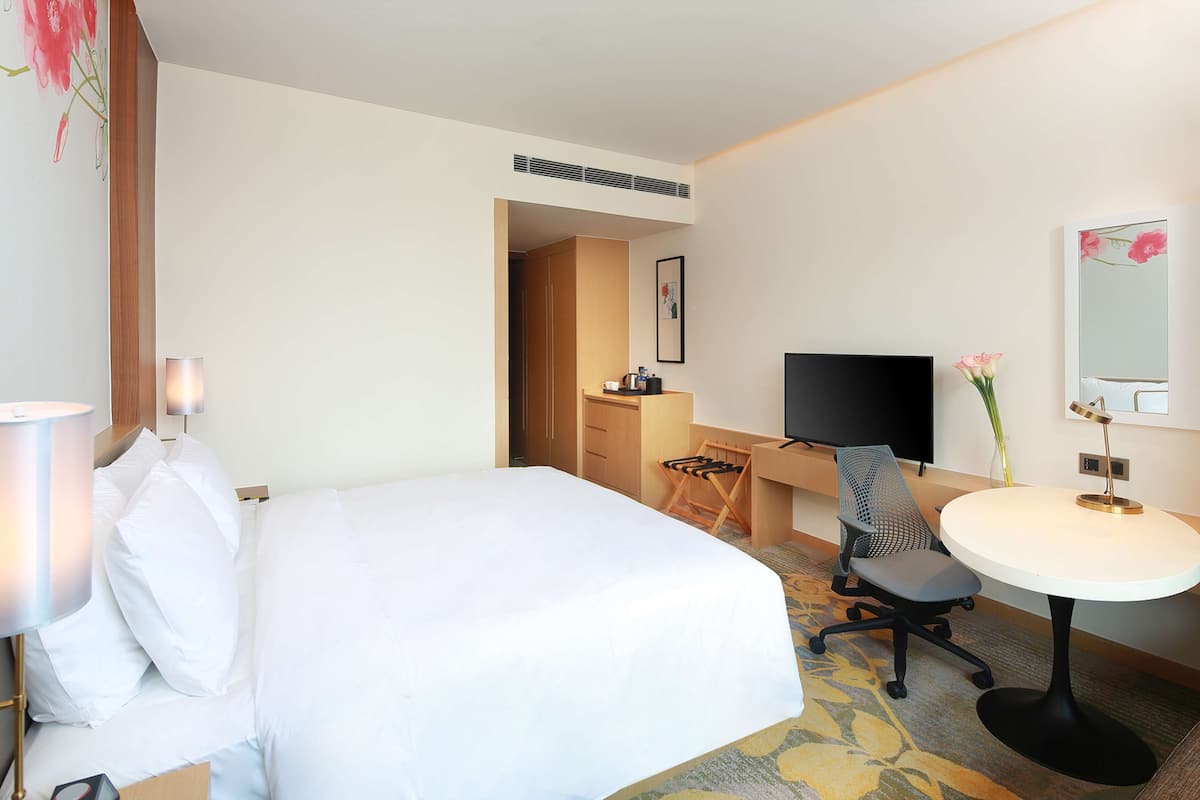 A hotel room with a bed, a desk with a chair, a TV, and a small table with a lamp.