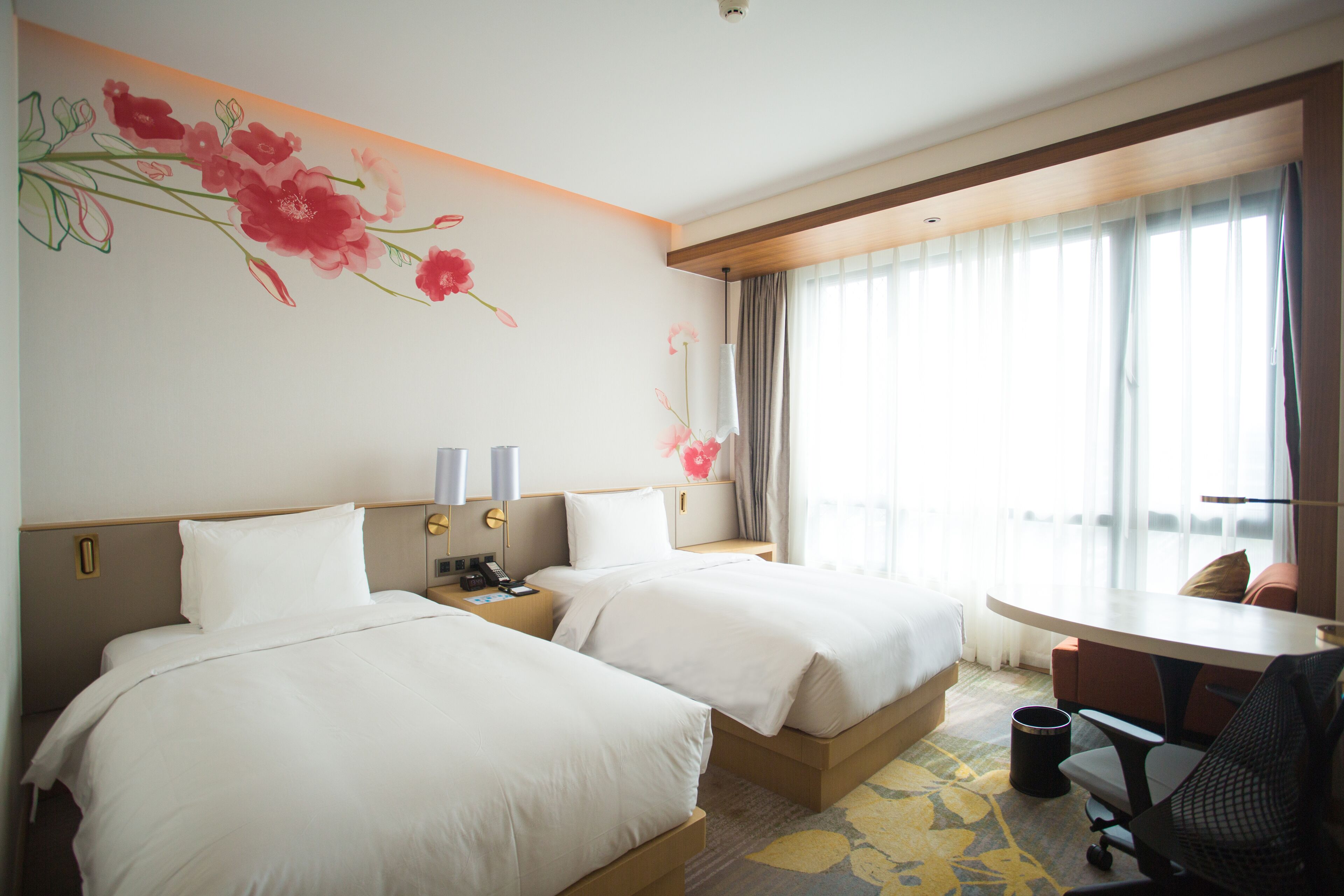 A hotel room with two beds, a desk, a chair, a trash bin, and a window with curtains.