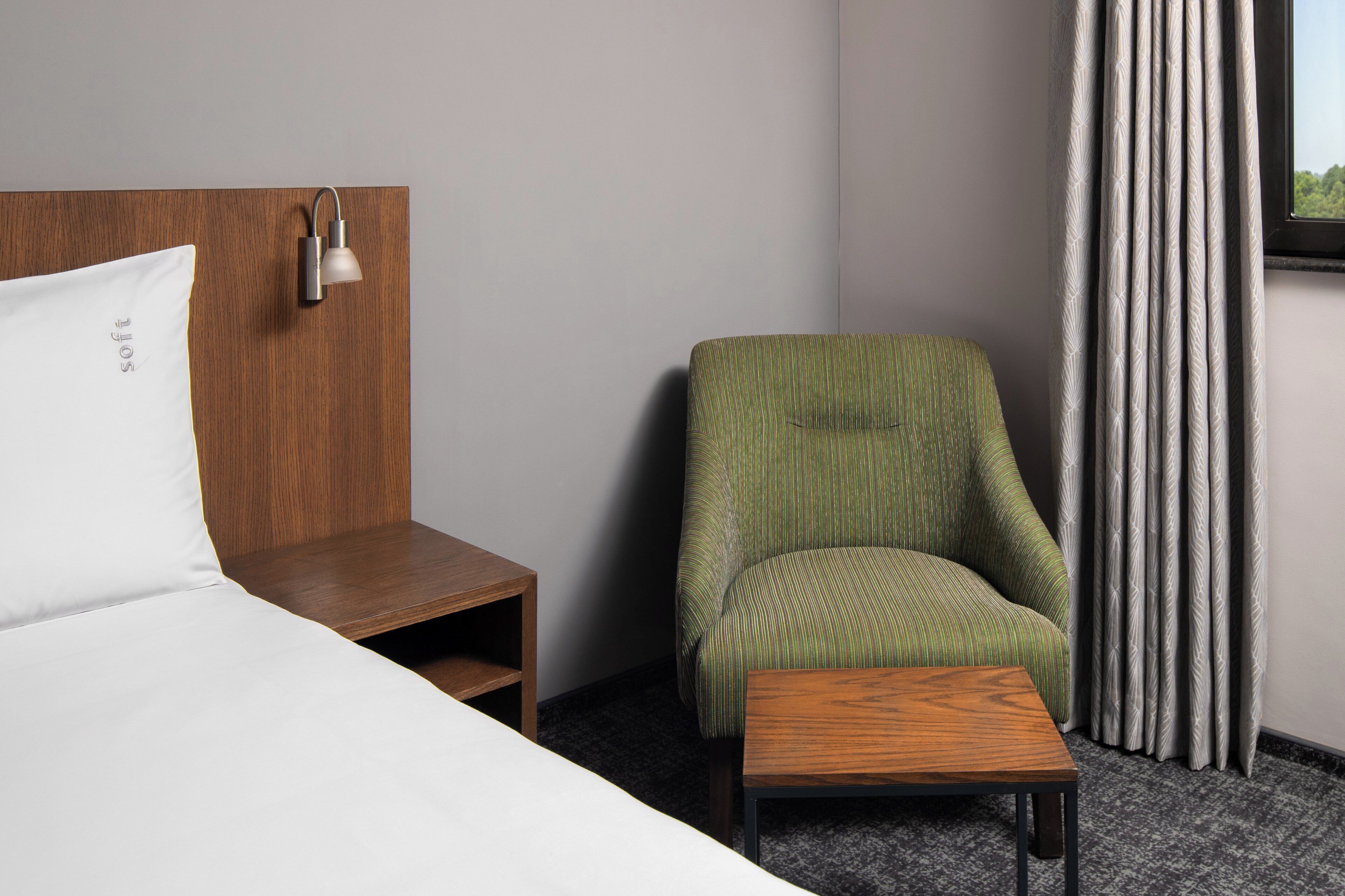 A hotel room with a bed, a nightstand, a green upholstered chair, a small wooden table, and a window with curtains.