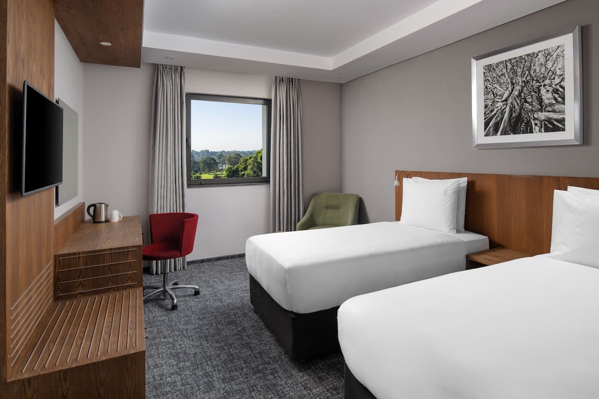 A hotel room with two beds, a desk with a flat-screen TV, a red chair, and a window with a view of trees.
