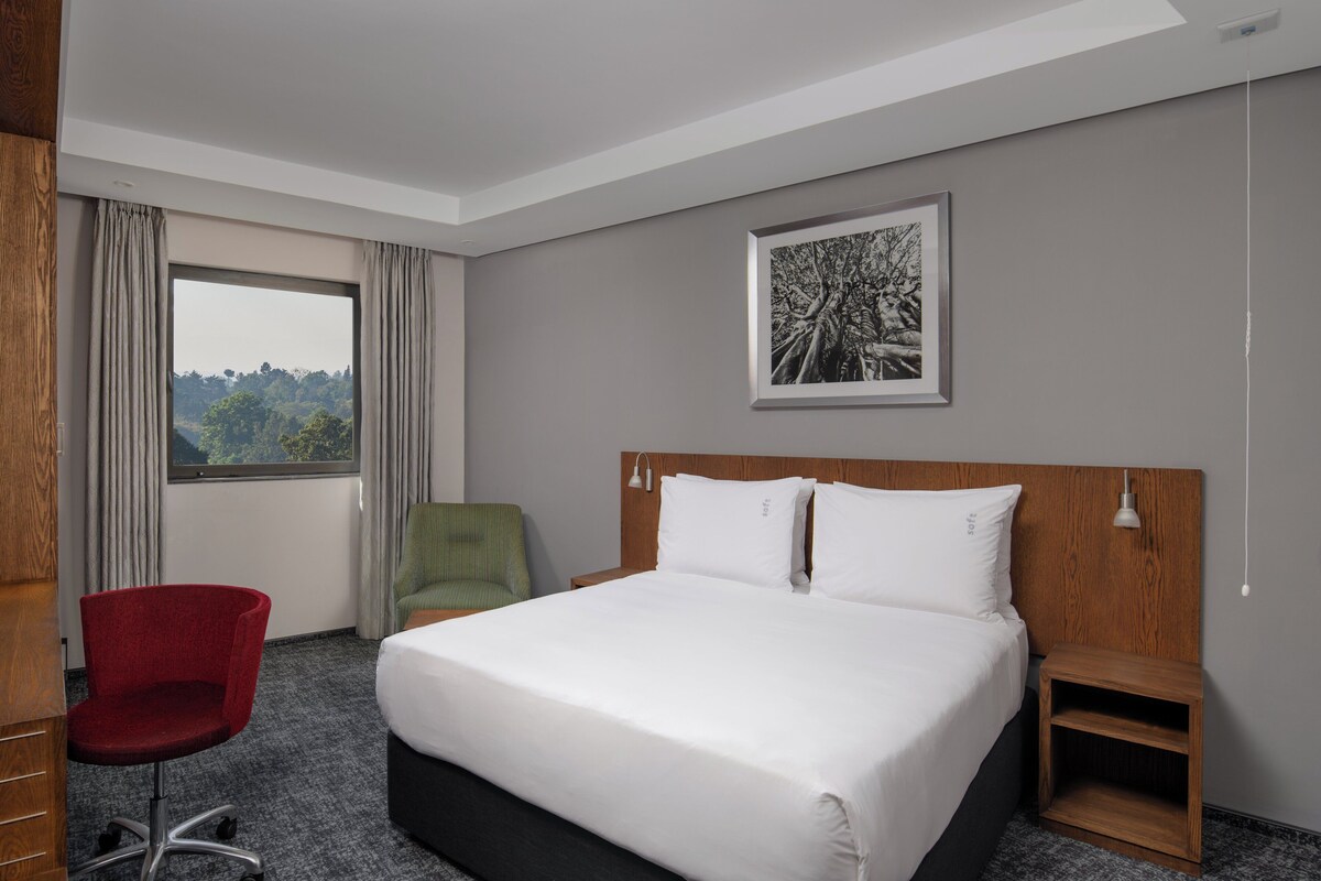 A hotel room with a large bed, a desk, a chair, a nightstand, and a window with curtains.