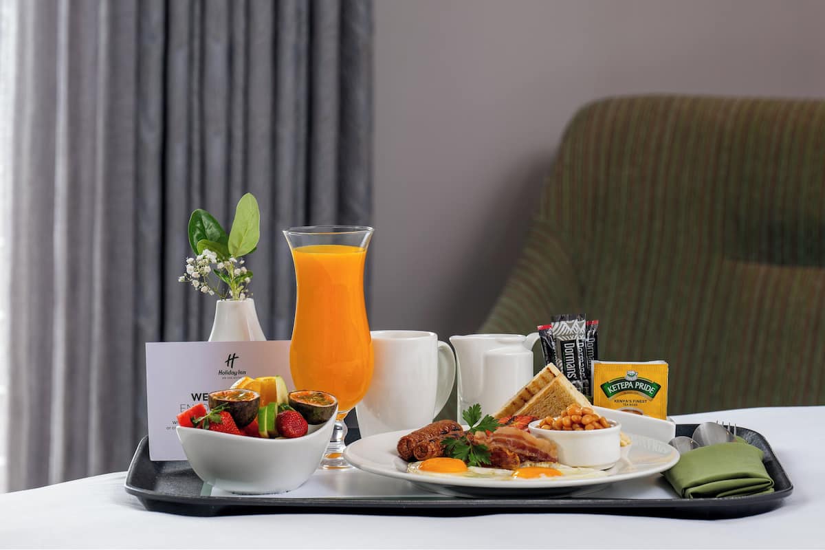 Una bandeja de desayuno con diversos alimentos, un vaso de zumo de naranja, un florero con flores y una tarjeta sobre una mesa.