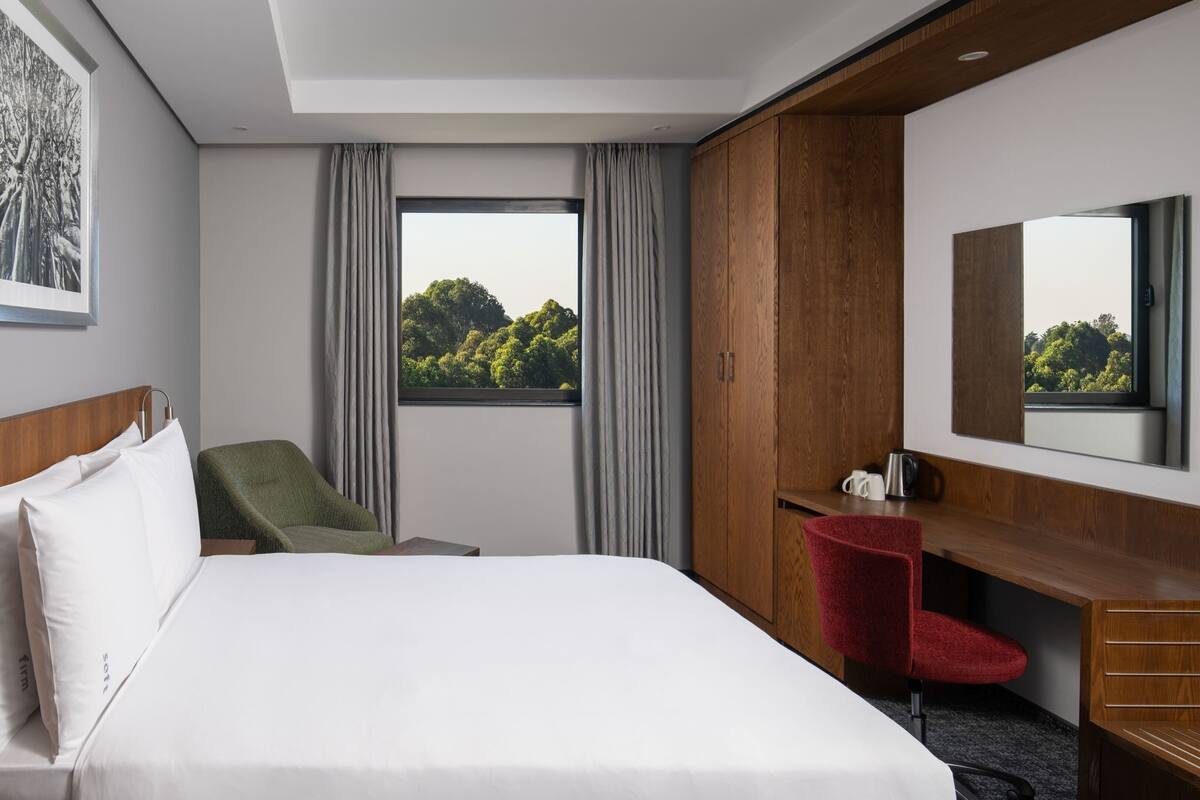 A hotel room with a large bed, a desk with a chair, a mirror, and a window with a view of trees.