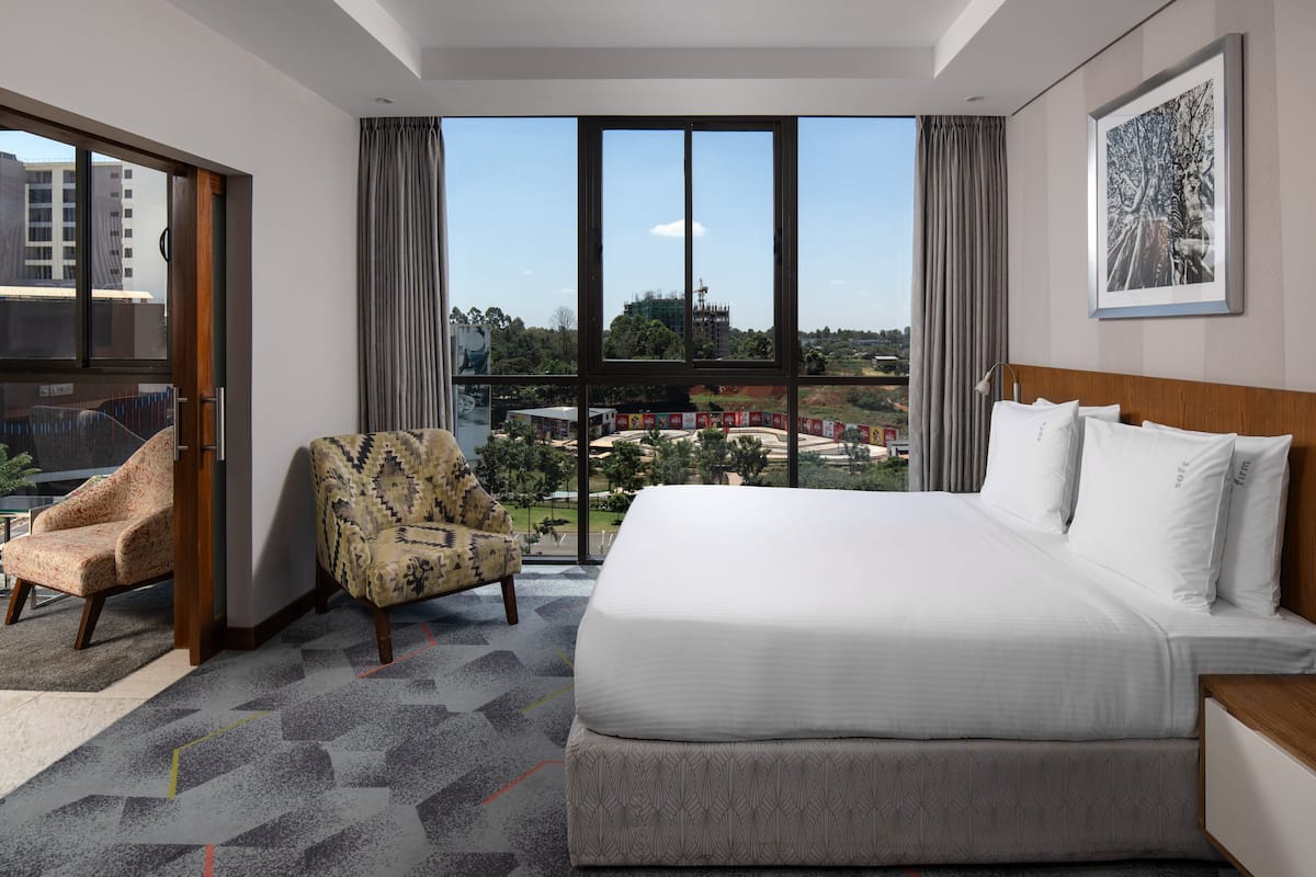 A hotel room with a large bed, two patterned armchairs, a view of a cityscape, and a framed picture on the wall.