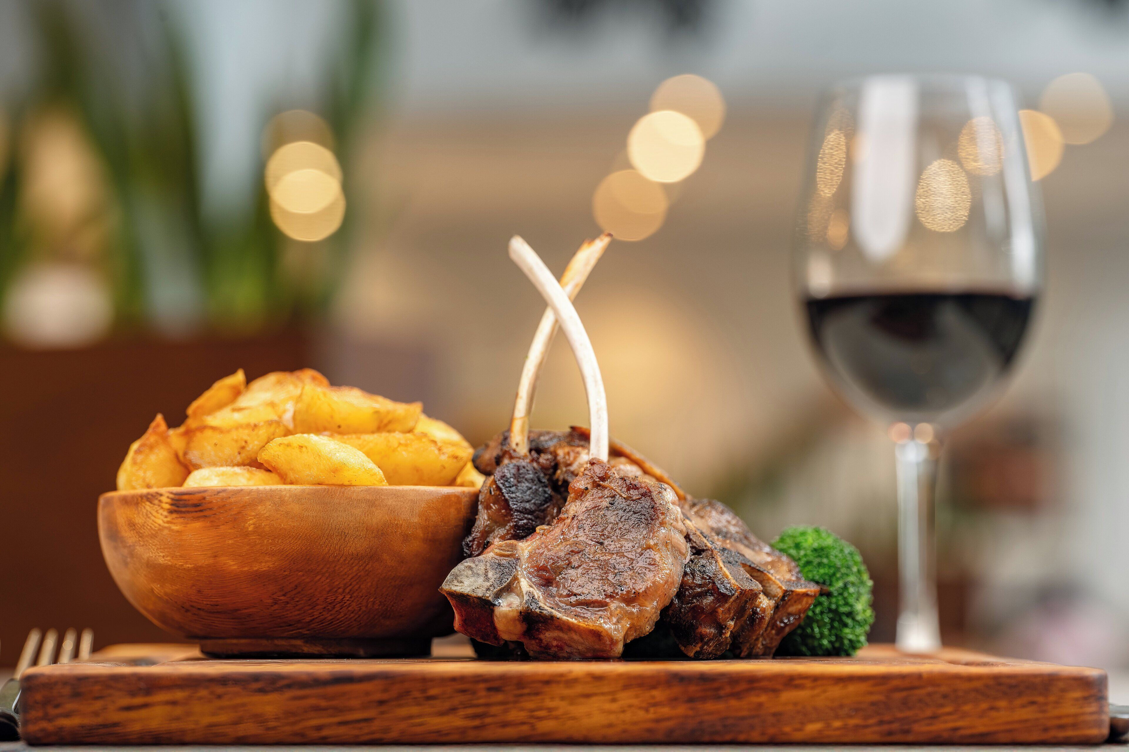 A wooden serving board with a bowl of potato wedges and a piece of meat, possibly lamb, with a glass of red wine.