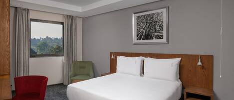 Habitación de hotel con una cama grande, una silla roja, una mesita de noche de madera y vistas al exterior a través de una ventana.