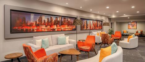 A modern lobby with a cityscape painting, a white sofa, orange and white armchairs, and a wooden coffee table.