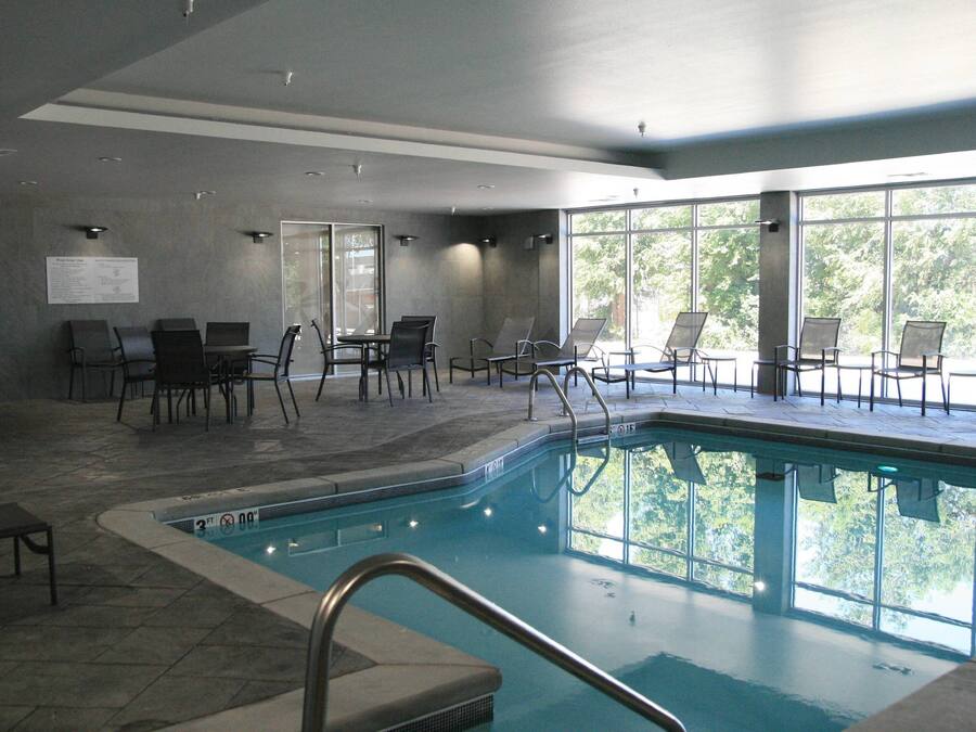 An indoor swimming pool with clear glass walls, surrounded by lounge chairs and tables.