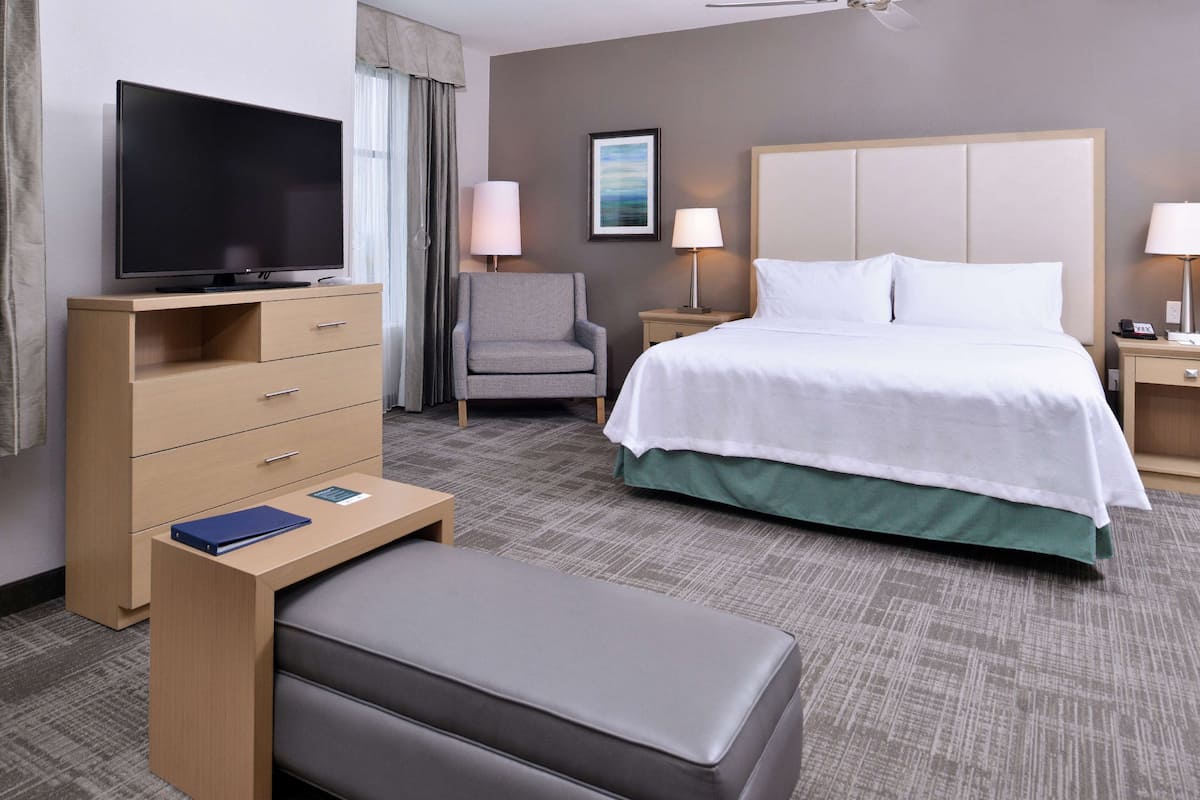 Habitación de hotel con cama, televisión sobre una cómoda de madera, una silla, dos lámparas de noche y una mesita con un libro.