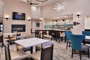 A hotel dining area with tables and chairs, a TV mounted on the wall, and a kitchen in the background. - Homewood Suites by Hilton Des Moines Airport (Des Moines)