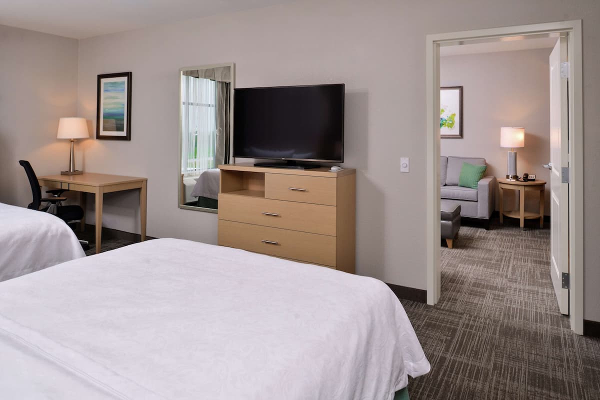 Habitación de hotel con dos camas, un escritorio con silla, televisor en una cómoda, un espejo y una mesita de noche con lámpara.