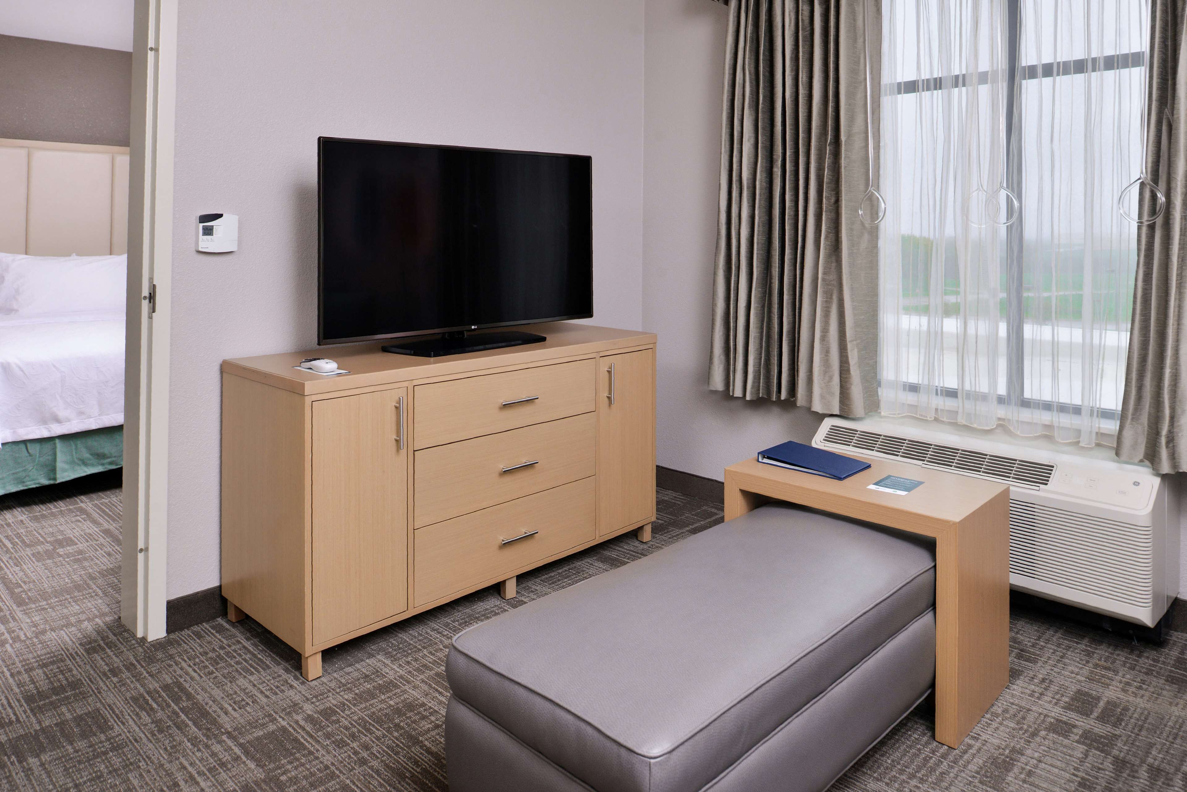 Hotelkamer met een bed, een televisie op een houten kast, een klein tafeltje met een boek en een afstandsbediening, en een raam met gordijnen.