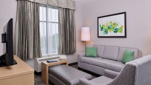 A hotel room with a grey sofa, a matching ottoman, a flat-screen TV on a wooden desk, a standing lamp, and a framed abstract painting on the wall.