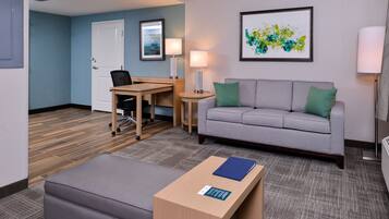 A hotel room with a grey sofa, a small table with a blue folder, a desk with a chair, a lamp, and a framed abstract painting on the wall.