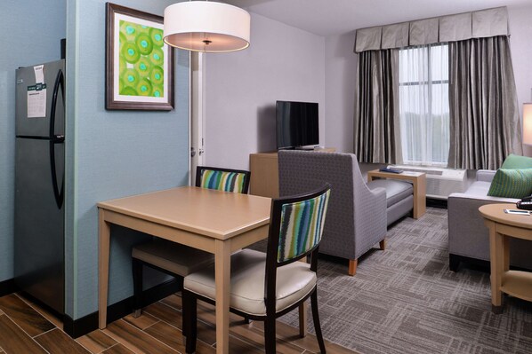 Habitación de hotel con mesa de comedor, dos sillas, un sofá, televisor y ventana con cortinas.