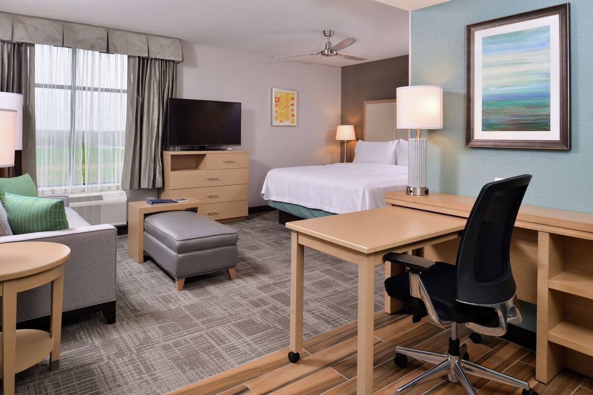 Habitación de hotel con cama, escritorio, silla, televisor y ventana con cortinas.