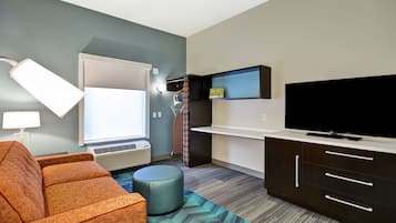 A modern hotel room with a sofa, ottoman, TV, and a small shelf.