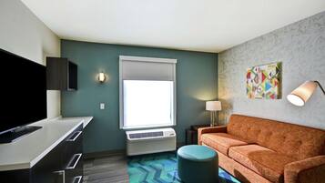 Una camera d'albergo moderna con una televisione a schermo piatto, un divano, un poggiapiedi, una finestra con tende e una lampada a parete, oltre a un quadro astratto dai colori vivaci.