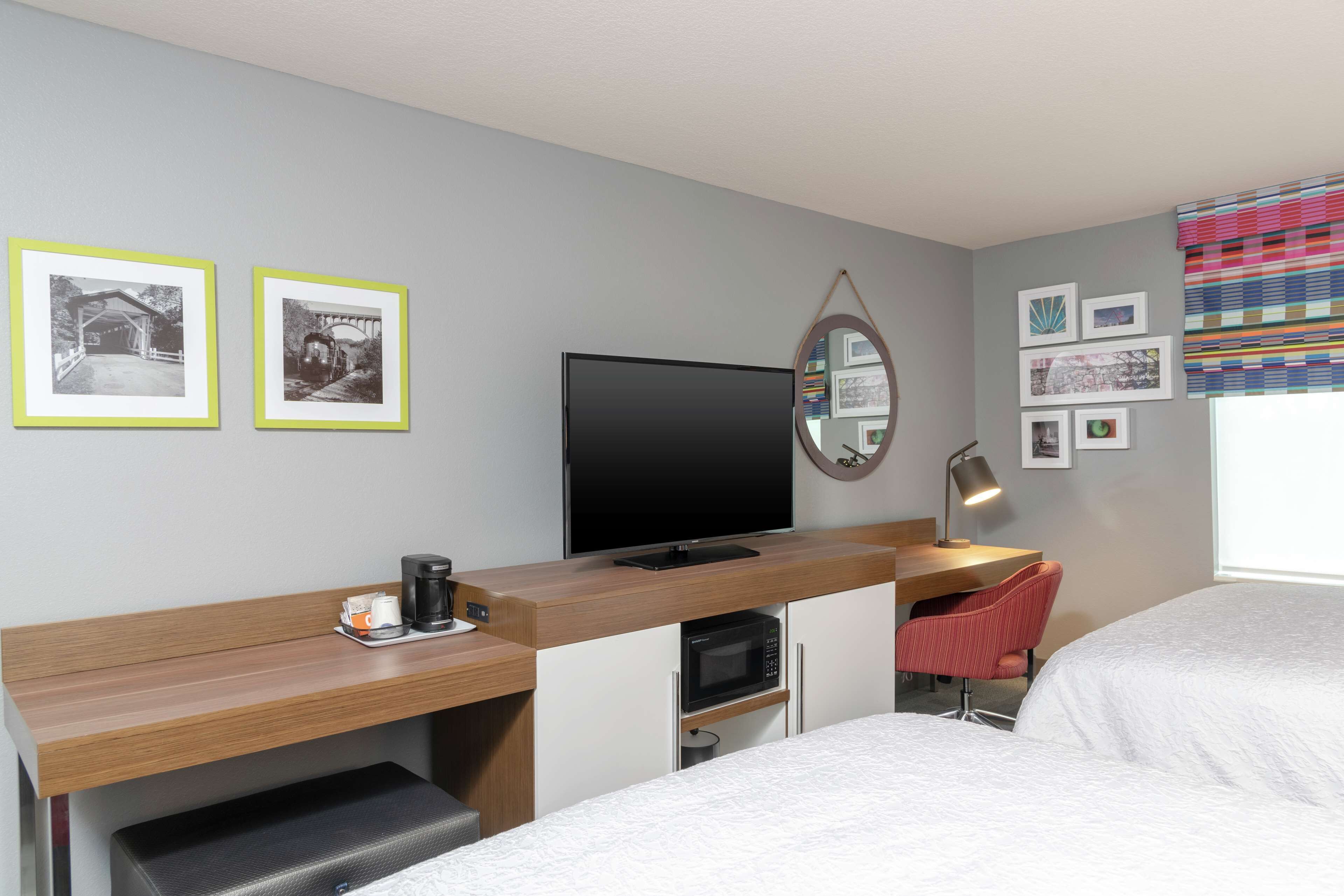 A hotel room with a bed, a desk with a flat-screen TV, a microwave, a coffee maker, and a chair. There are framed pictures on the wall.