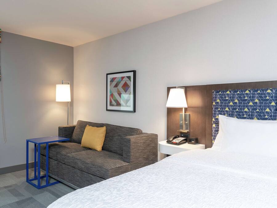 A hotel room with a bed, a sofa, a small table, a lamp, a phone, and a wall-mounted artwork.