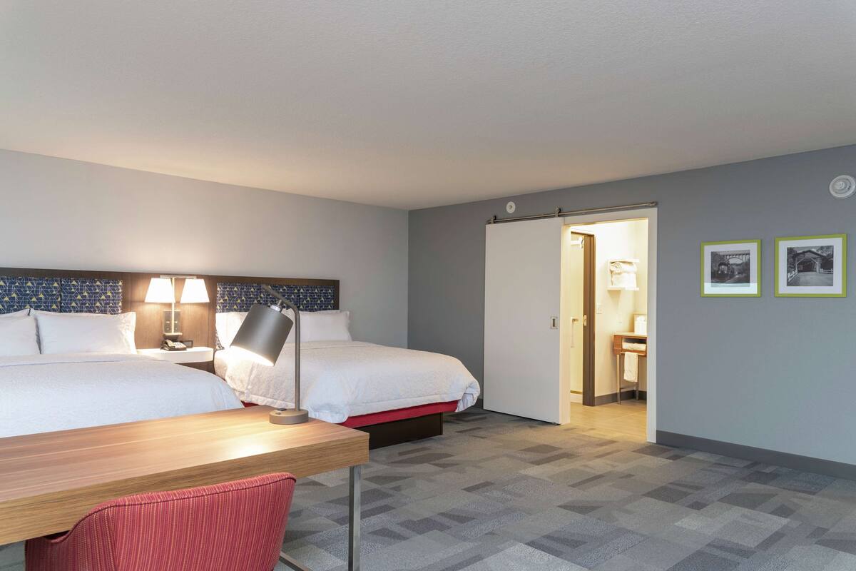 A hotel room with two beds, a desk, a chair, and a bathroom visible through an open door.