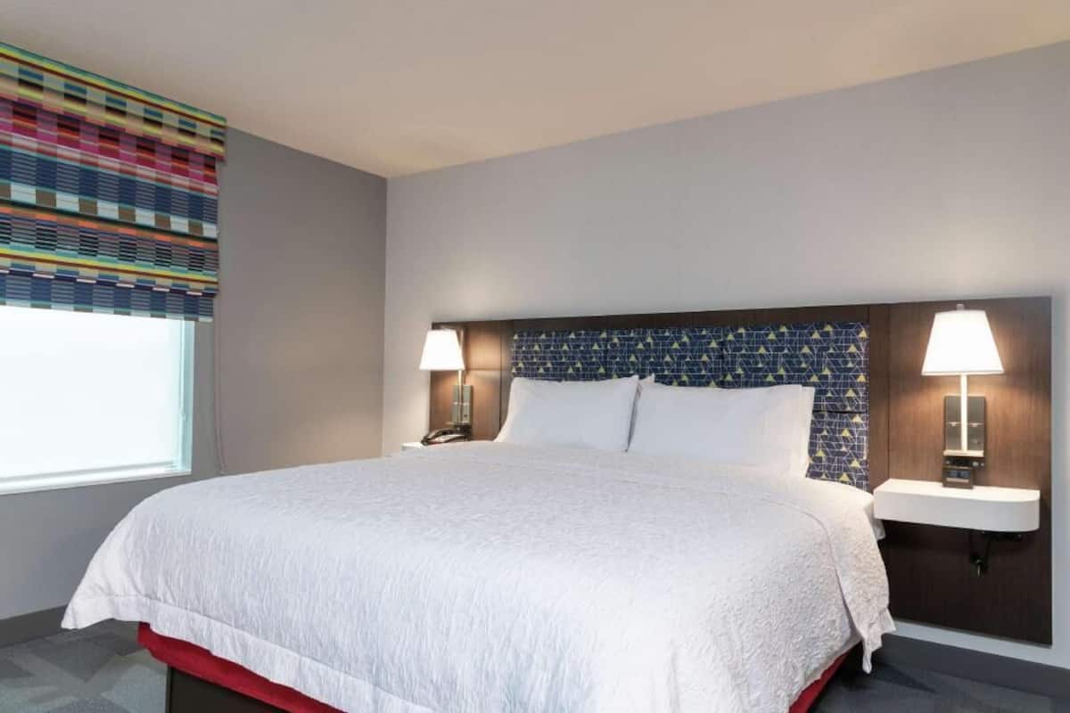 A hotel room with a large bed, two bedside lamps, a nightstand, a window with blinds, and a wall with a geometric pattern.