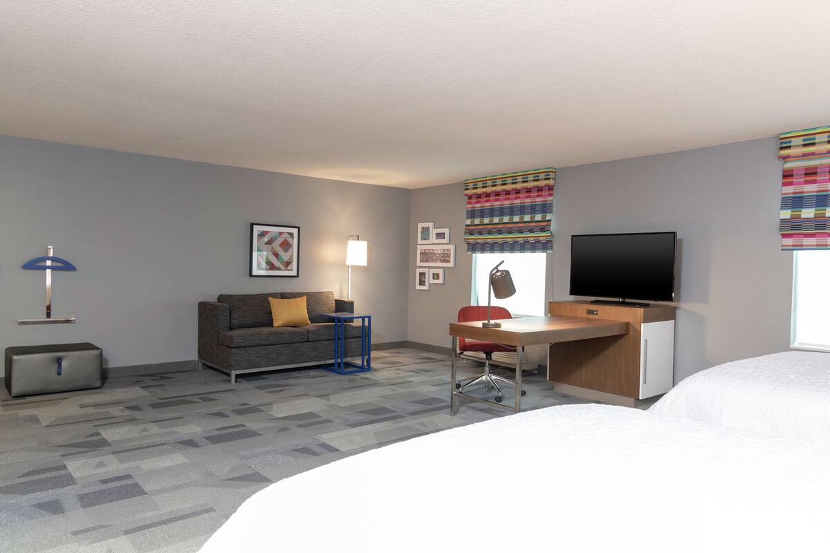 A hotel room with a bed, a desk, a television, a sofa, a lamp, and a suitcase.
