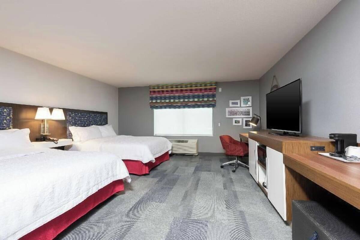 A hotel room with two beds, a desk, a television, and a window with a striped curtain.