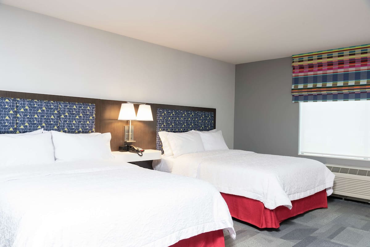 A hotel room with two beds, a nightstand with a lamp, a patterned headboard, and a window with a colorful curtain.