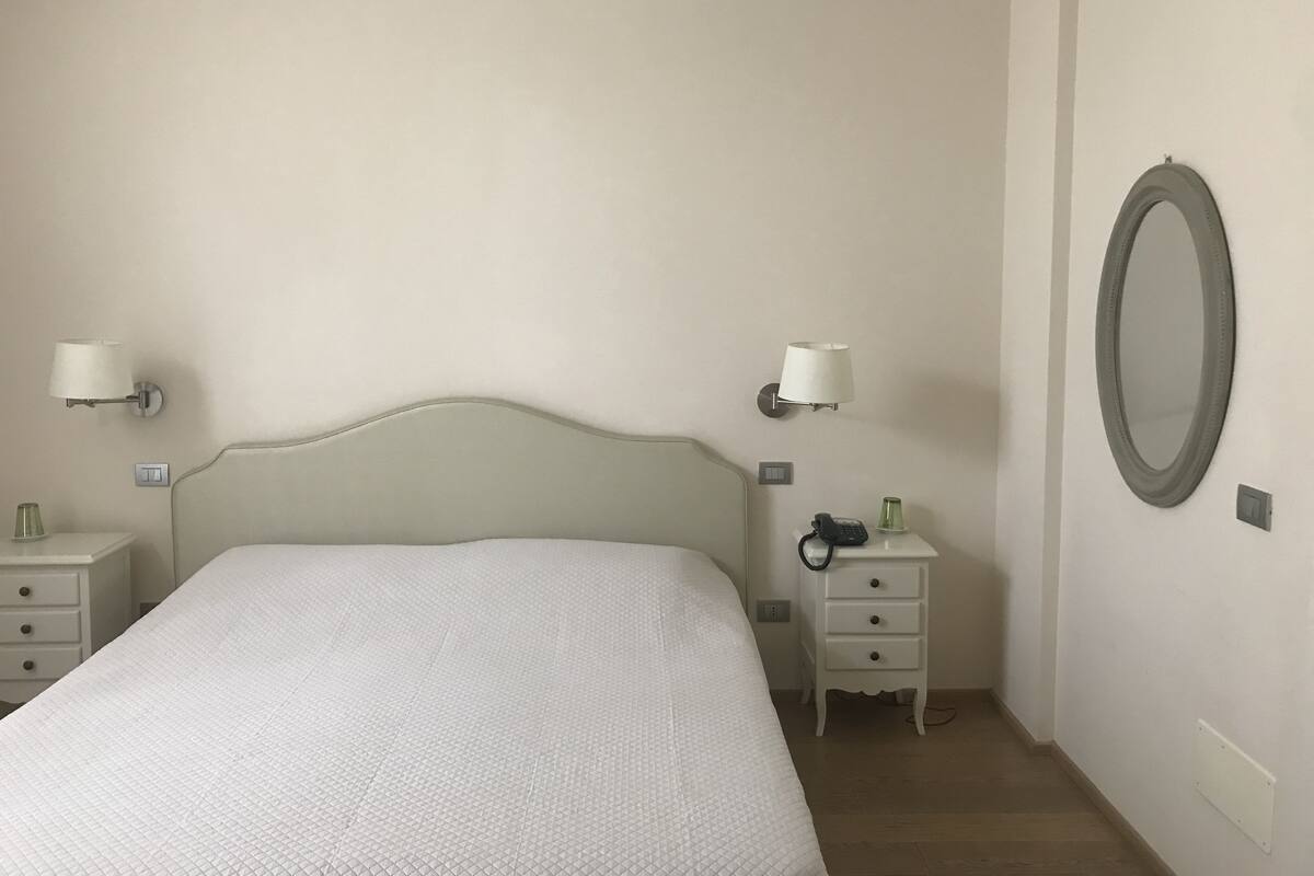 A bedroom with a bed, two bedside tables, a nightstand, a lamp, a telephone, and a mirror.