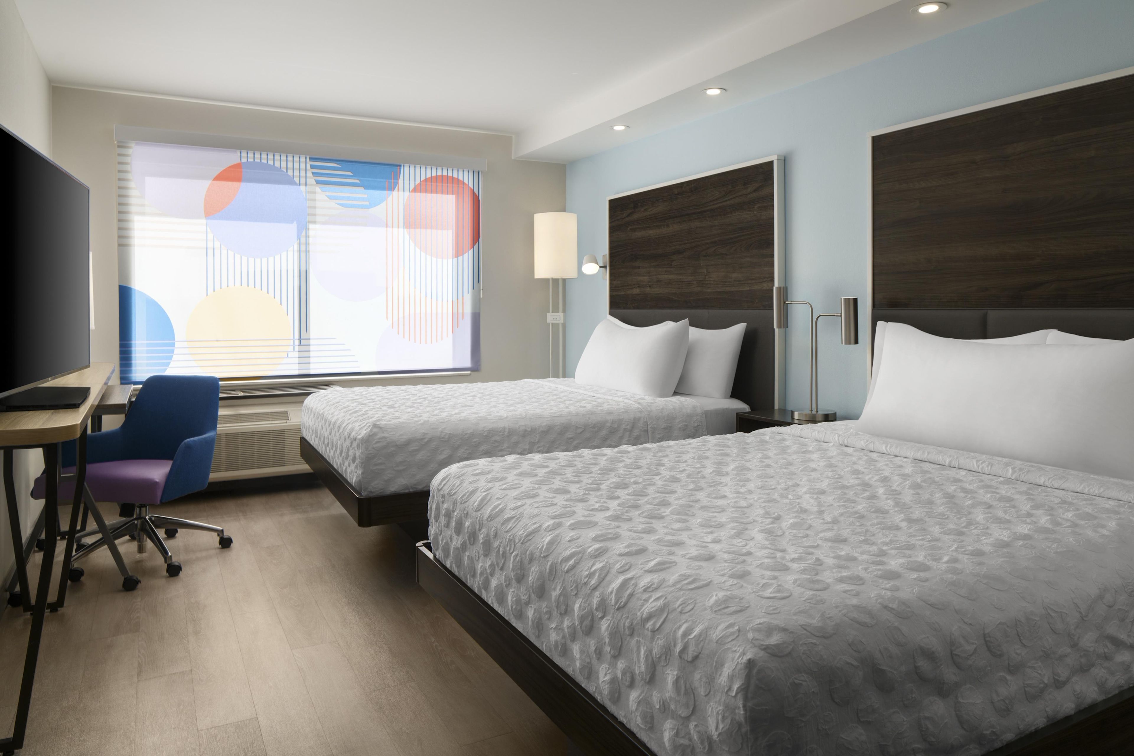 A hotel room with two beds, a desk, a chair, and a TV.