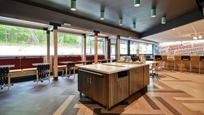 A modern restaurant interior with a central wooden bar, seating along the walls, and large windows offering a view of the outdoors. - Tru by Hilton Chicopee Springfield (Chicopee)
