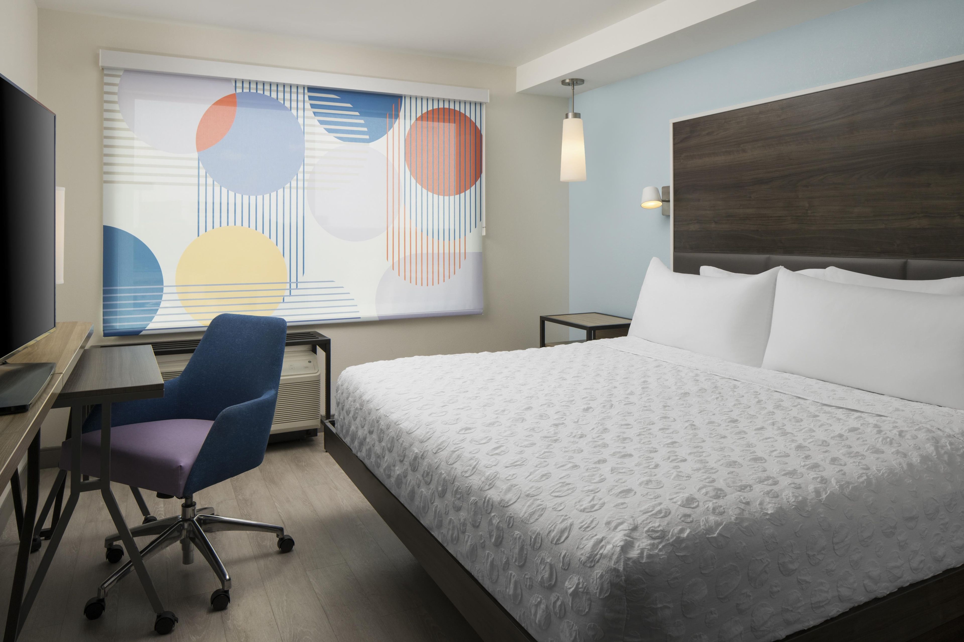 A hotel room with a bed, a desk, a chair, a TV, and a window with abstract art.