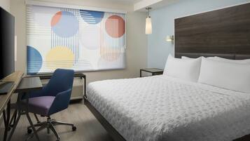 A hotel room with a bed, a desk, a chair, a TV, and a window with abstract art.