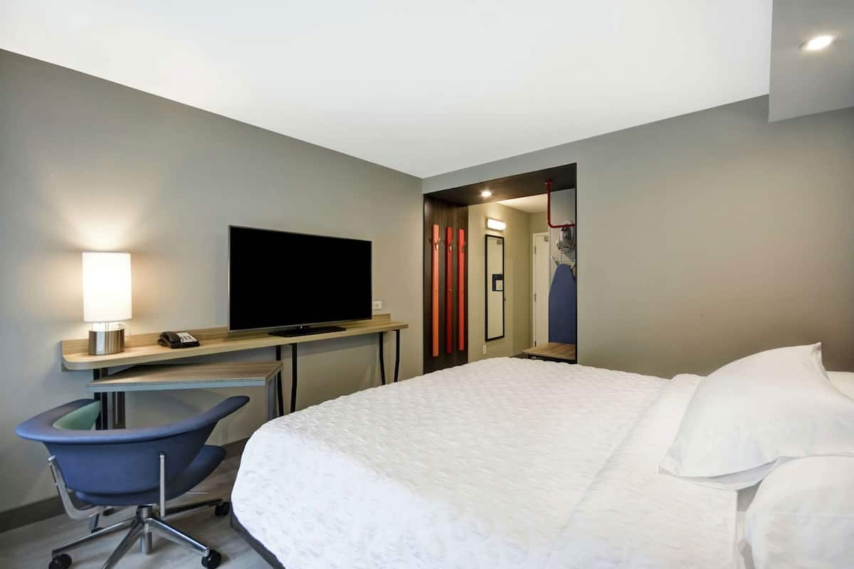 Una habitación de hotel con una cama, un escritorio con televisor, una silla y un espejo.